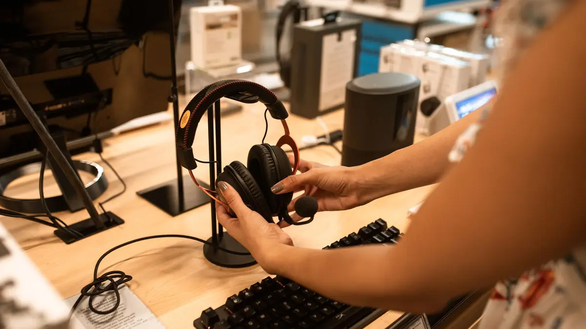 Deux mains ajustent un casque audio noir sur un support, sur le comptoir en bois clair d'une boutique d'électronique