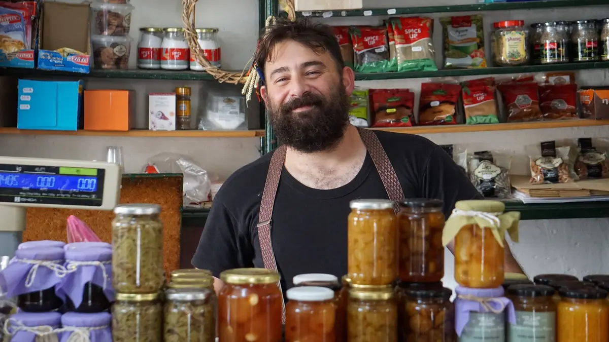 Un commerçant barbu en tablier sourit derrière un comptoir de bocaux de confiture, devant des étagères d'épices