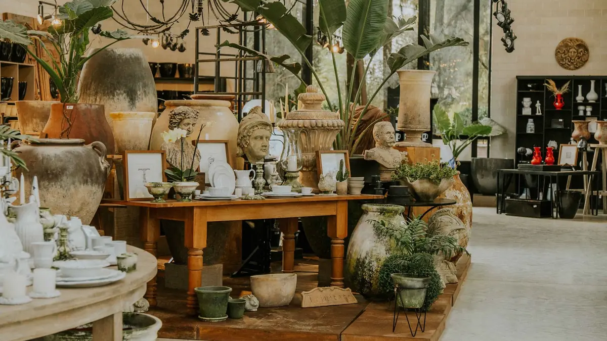 Intérieur d'une boutique de poterie et de déco : table en bois avec vaisselle blanche, grandes jarres en terre cuite, plantes et lustre