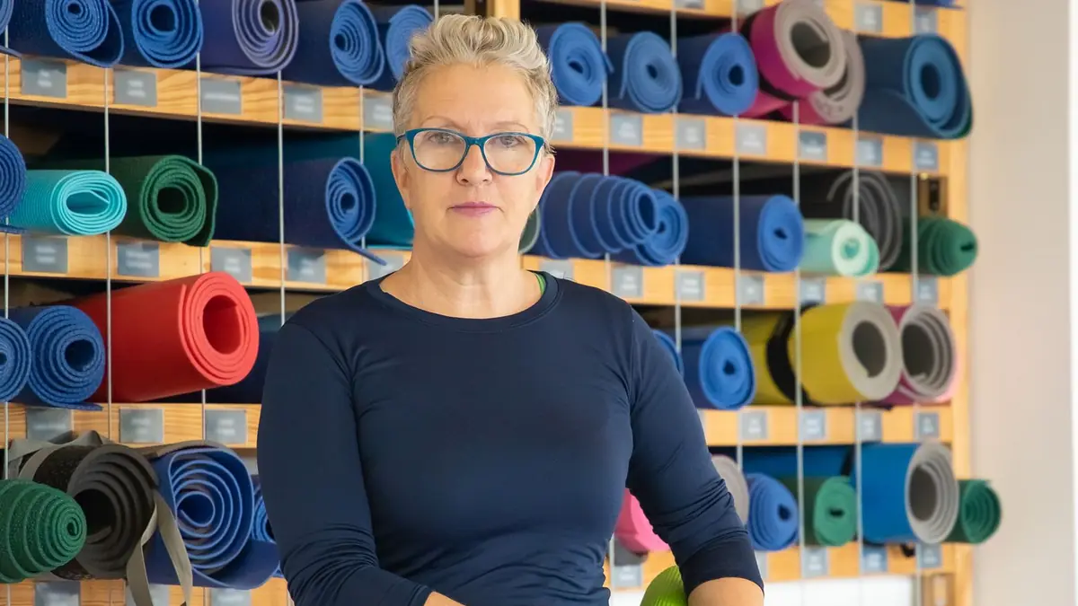 Une femme aux cheveux gris et lunettes bleues tient un tapis de yoga vert devant un mur de tapis roulés multicolores